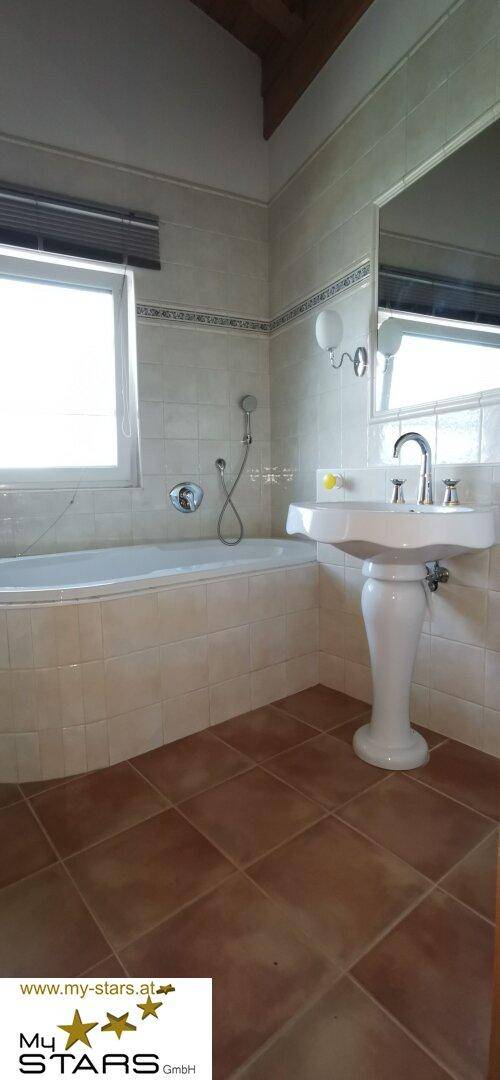 Badezimmer mit Badewanne, Waschbecken und Fenster, mit braunen Fliesen.