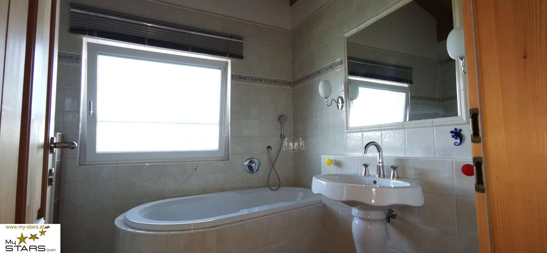 Badezimmer mit Badewanne, Waschbecken und Fenster, unter einer Holzbalkendecke.