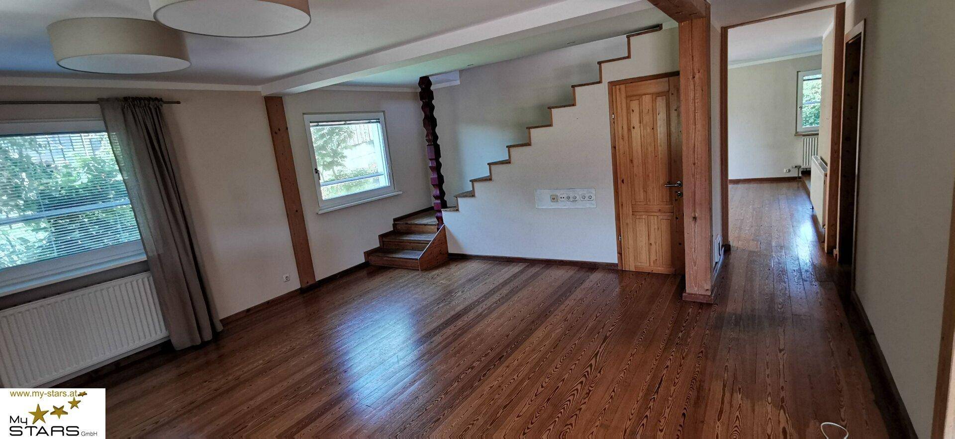 Helles Wohnzimmer mit Holzboden, Treppe und Fenstern, die viel Tageslicht hereinlassen.
