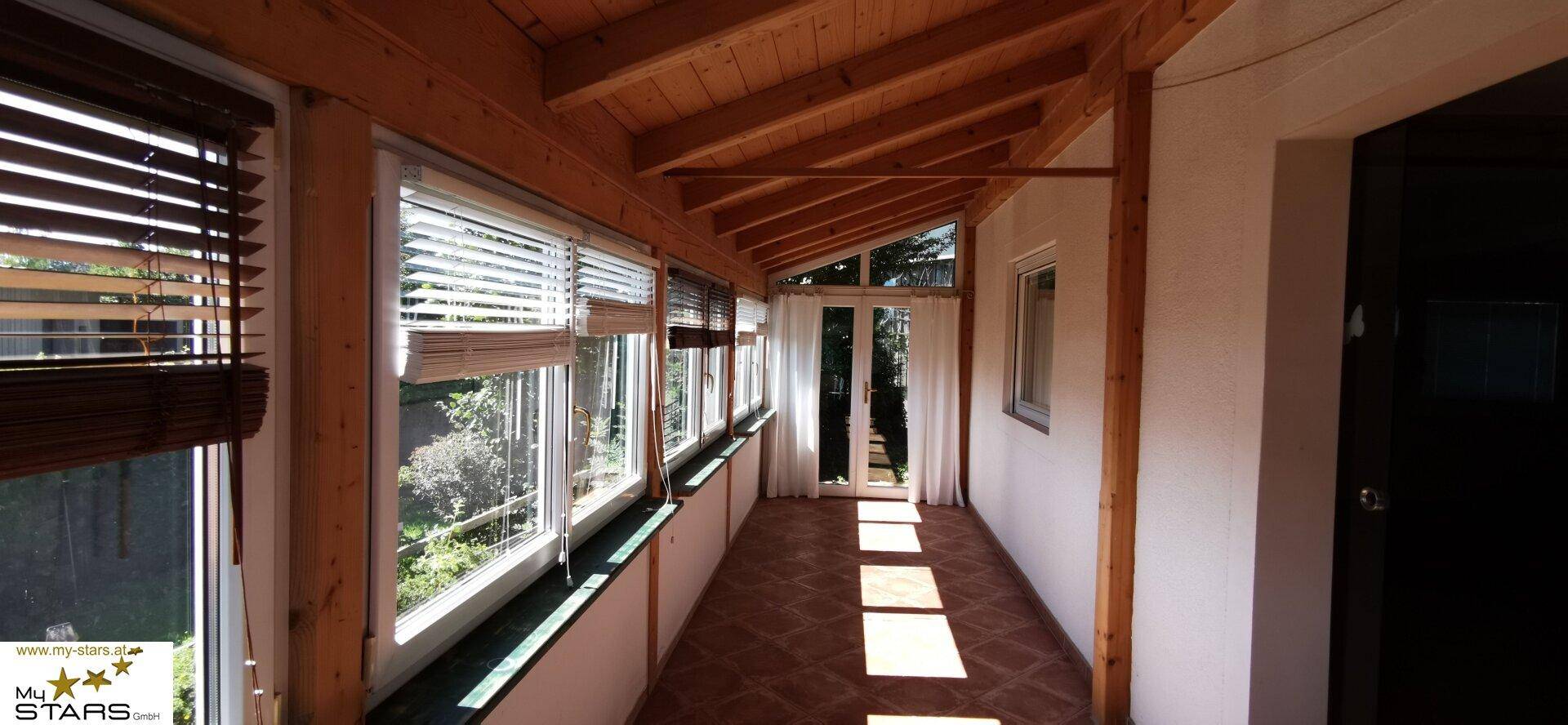 Heller Wintergarten mit vielen Fenstern, Holzbalkendecke und Fliesenboden, ideal zum Entspannen.