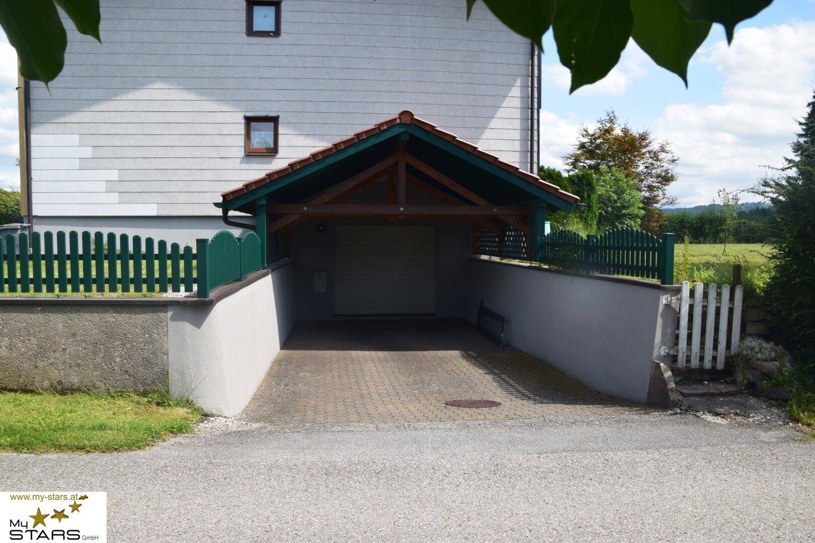 Überdachter Garagenzugang mit gepflasterter Einfahrt und grünem Holzzaun.