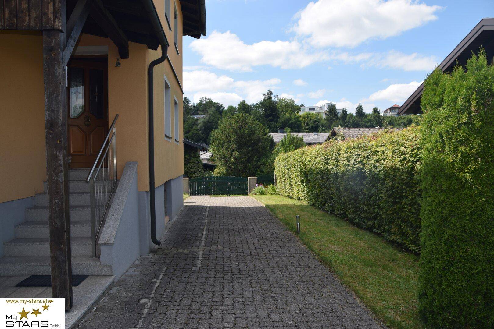 Gepflasterte Zufahrt zum Hauseingang, gesäumt von einer hohen Hecke und Bäumen.