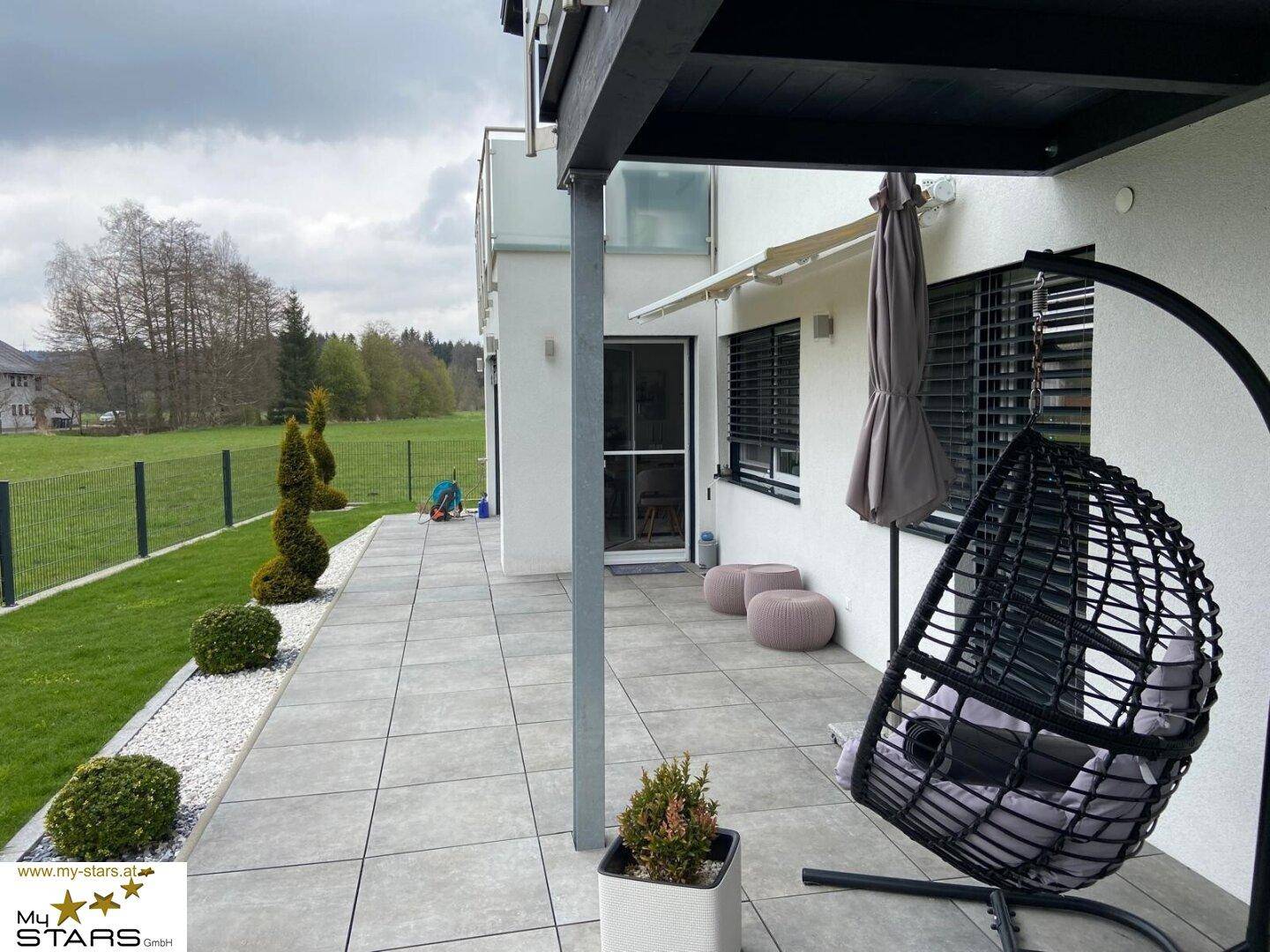 Gepflasterte Terrasse mit Hängesessel, Sonnenschirm und Blick auf den gepflegten Gartenbereich.