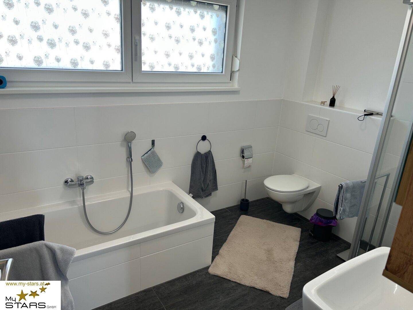 Modernes Badezimmer mit Badewanne, WC und Fenster, ausgestattet mit weißen und dunklen Fliesen.