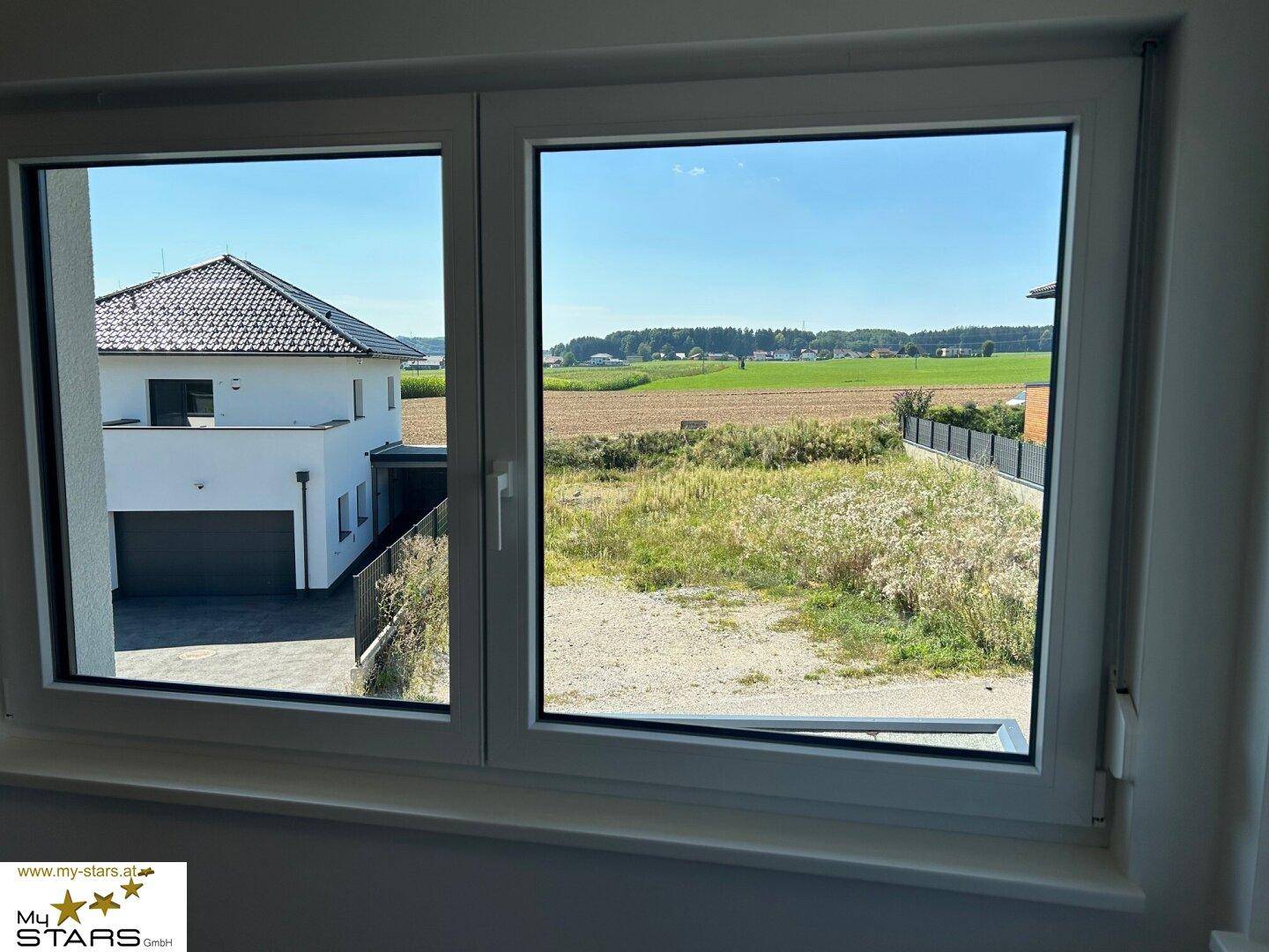Fensterblick auf die umliegende Landschaft mit einem Nachbarhaus und weiten Feldern.