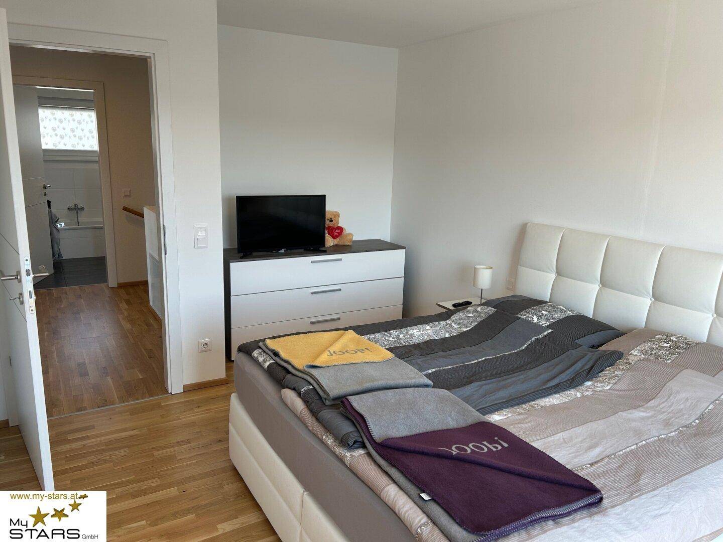 Helles Schlafzimmer mit Doppelbett und Kommode, angrenzend an einen Flur.