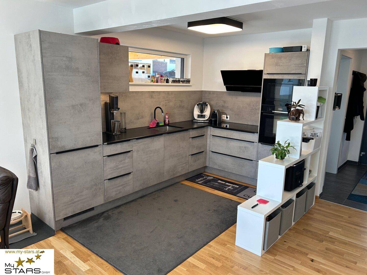 Moderne Küche mit grauen Fronten, schwarzen Arbeitsplatten und integrierten Geräten, offen zum Wohnbereich.