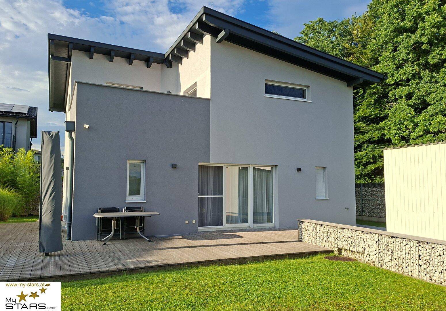Rückansicht des modernen Hauses mit großer Holzterrasse und angrenzendem grünen Garten.