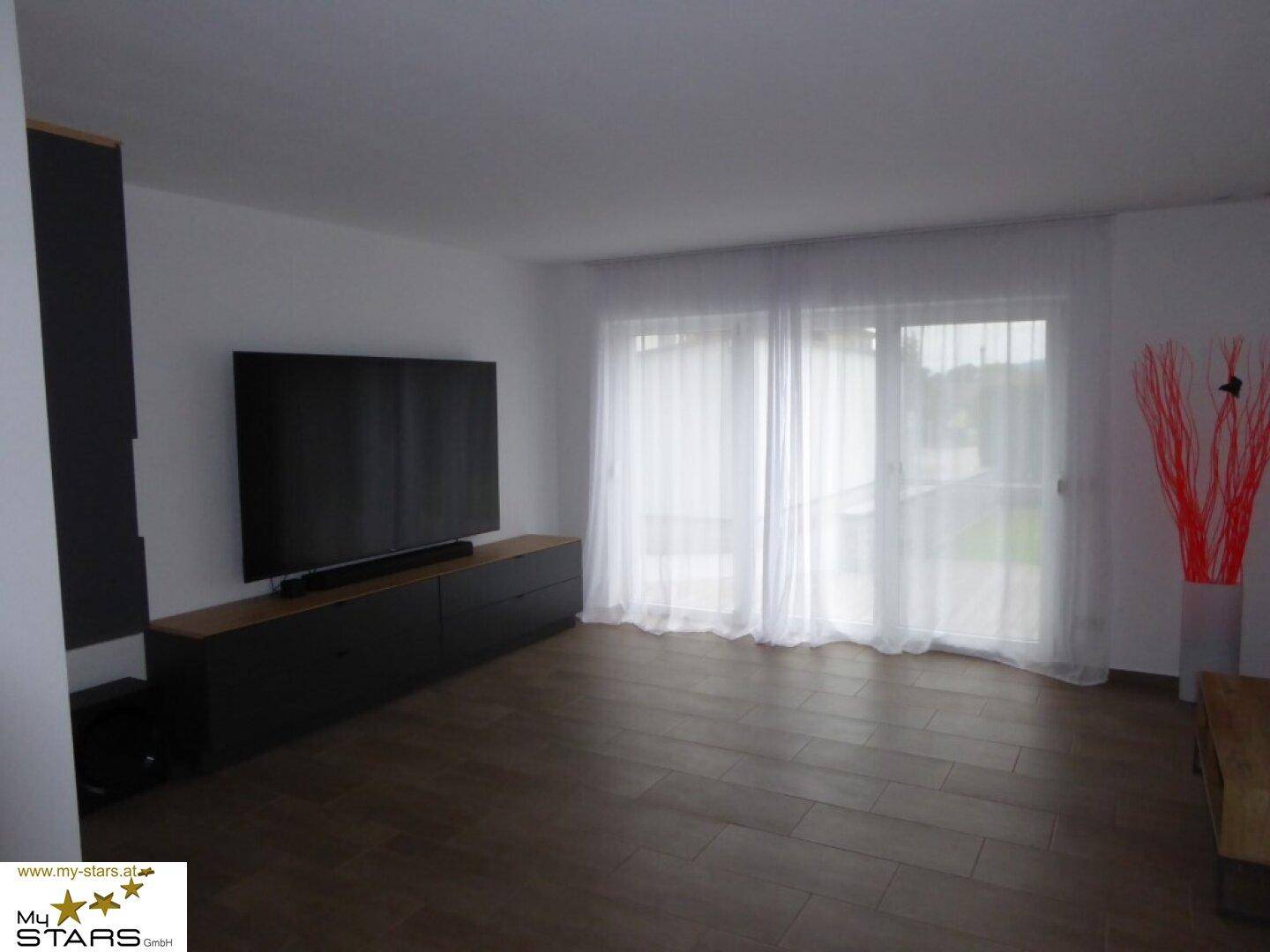 Geräumiges Wohnzimmer mit großem Fernseher, Medienmöbeln und viel Tageslicht.
