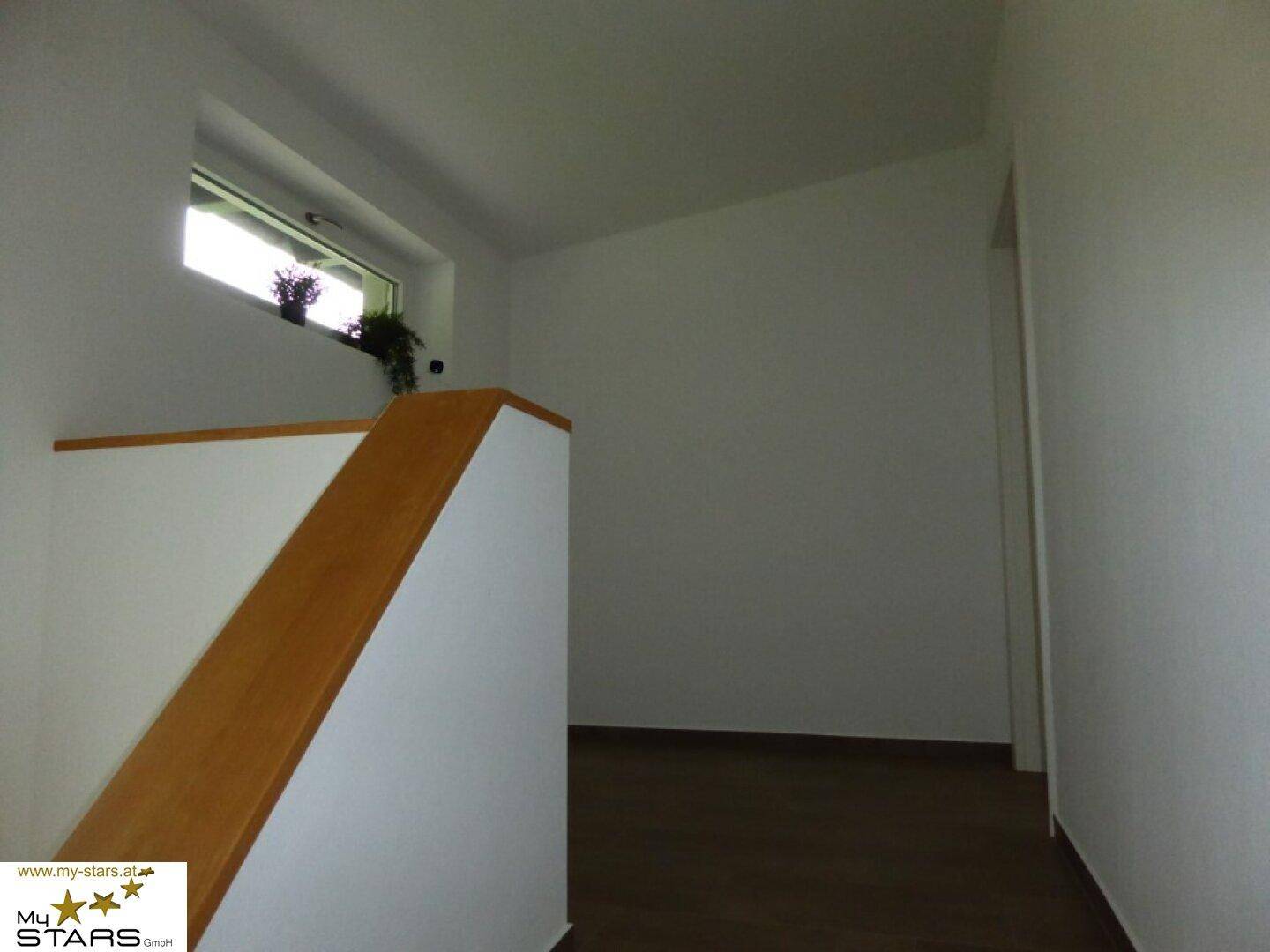 Heller Flur mit Holztreppengeländer und dunklem Laminatboden, der zu weiteren Räumen führt.