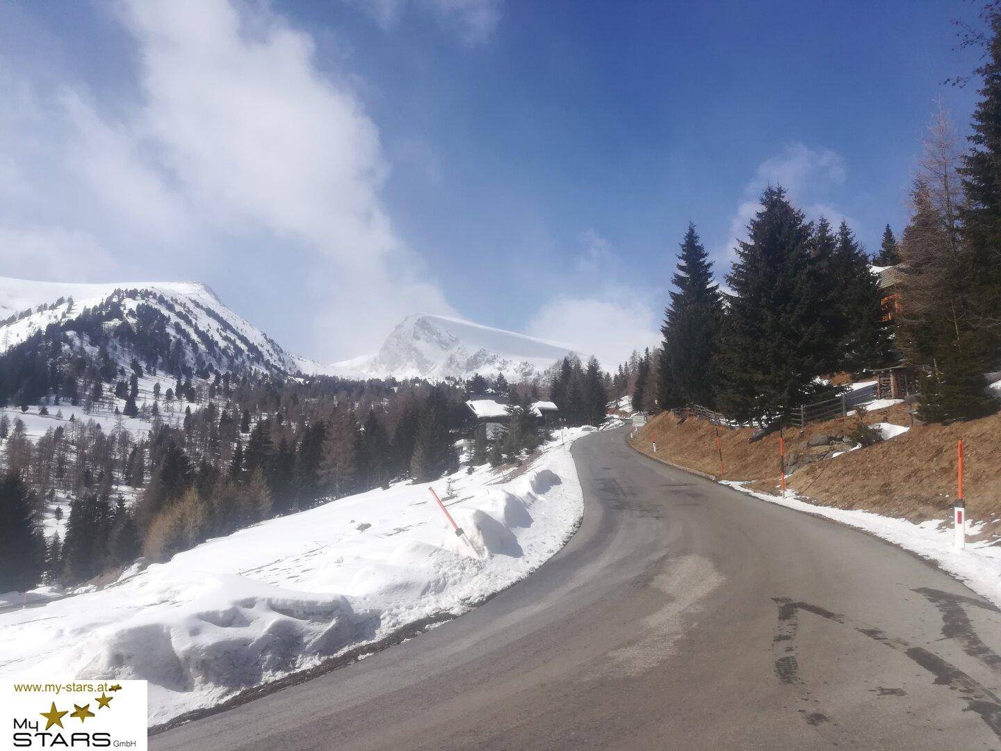 Eine verschneite Bergstraße führt durch eine Winterlandschaft mit Kiefern und schneebedeckten Gipfeln.