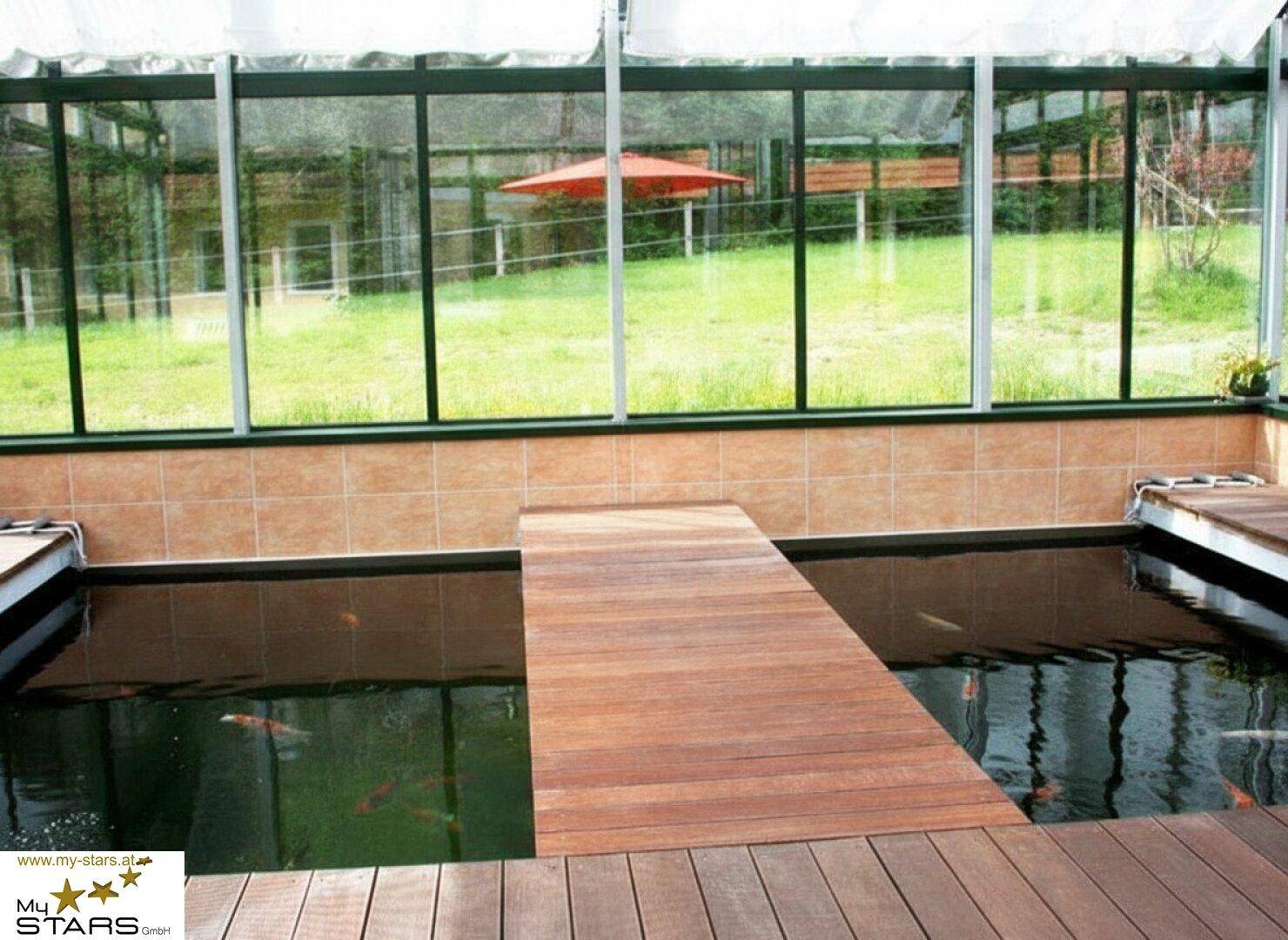 Heller Wintergarten mit großem Fischteich und Holzsteg, umgeben von Glaswänden und Blick ins Grüne.