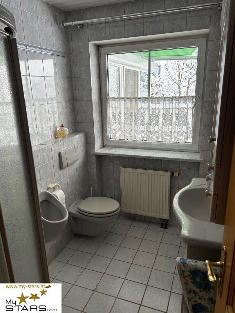 Kleines Badezimmer mit Fliesen, WC, Urinal, Dusche und Fenster.