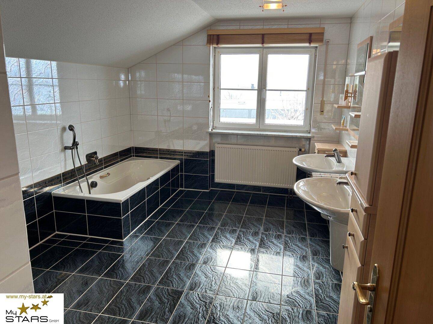 Badezimmer mit dunklen Fliesen, Badewanne und Doppelwaschbecken unter Dachschräge.