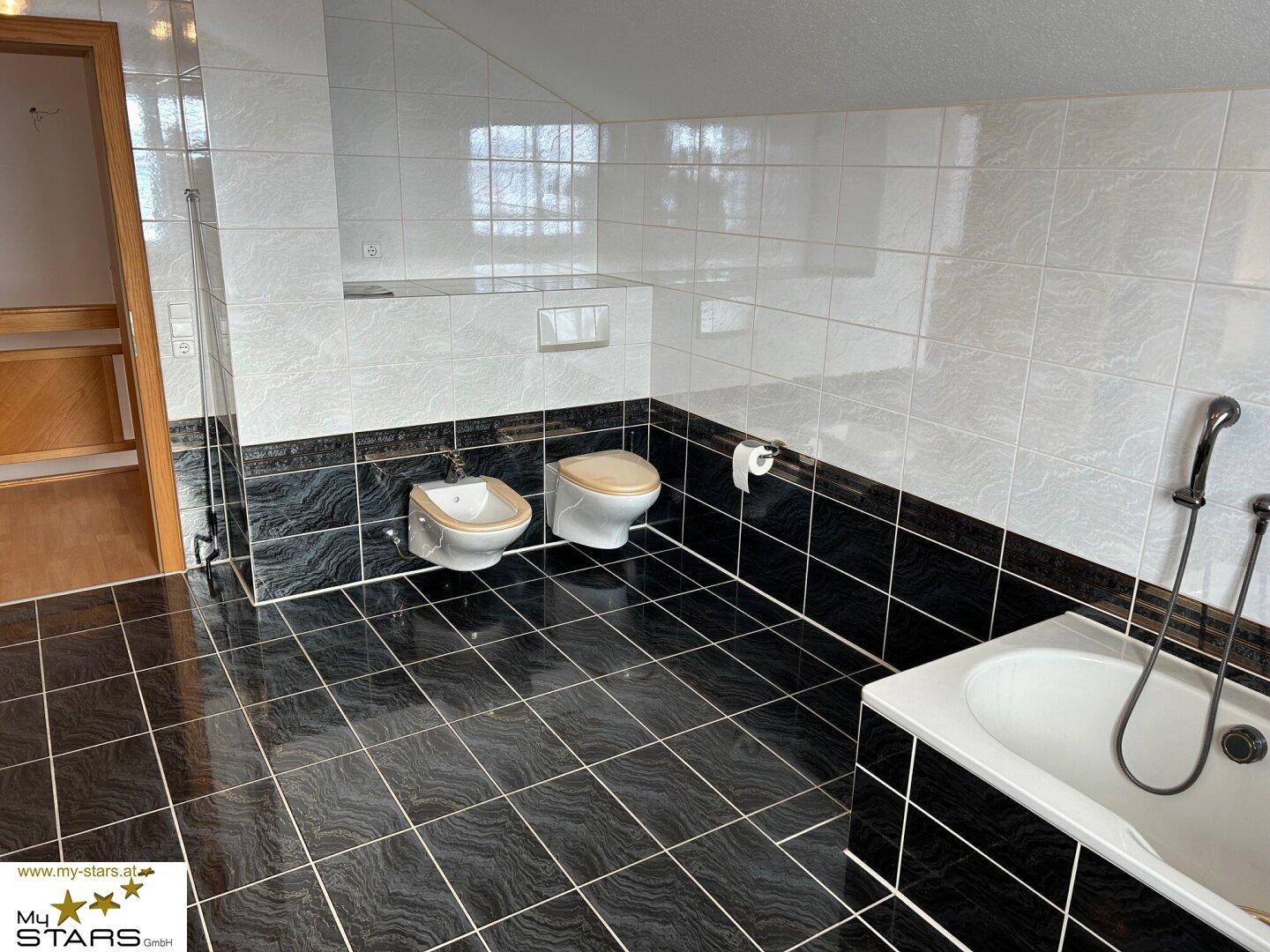 Badezimmer mit Badewanne, Bidet, WC und dunklen Fliesen unter Dachschräge.