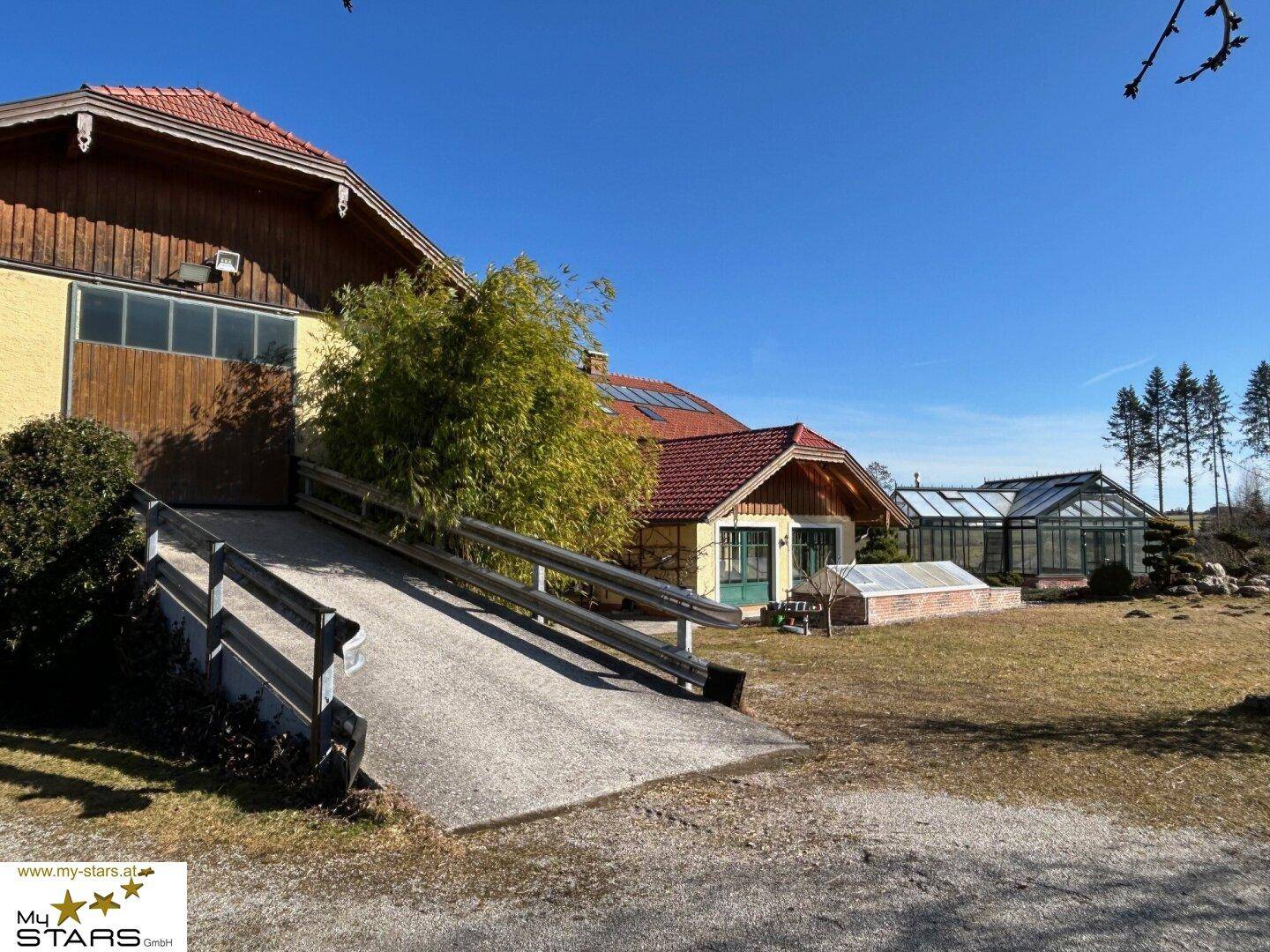 Gebäudefassade mit Zufahrtsrampe und angrenzendem Gartenbereich unter strahlend blauem Himmel.
