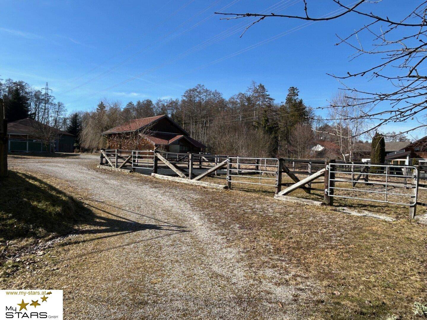 Schotterweg mit Holzzäunen und Bäumen, der zu weiteren Gebäuden des Anwesens führt.