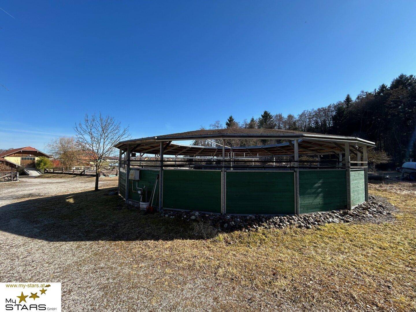 Rundes Reitgebäude mit offenem Dach und grünen Seitenwänden, eingebettet in die Landschaft.