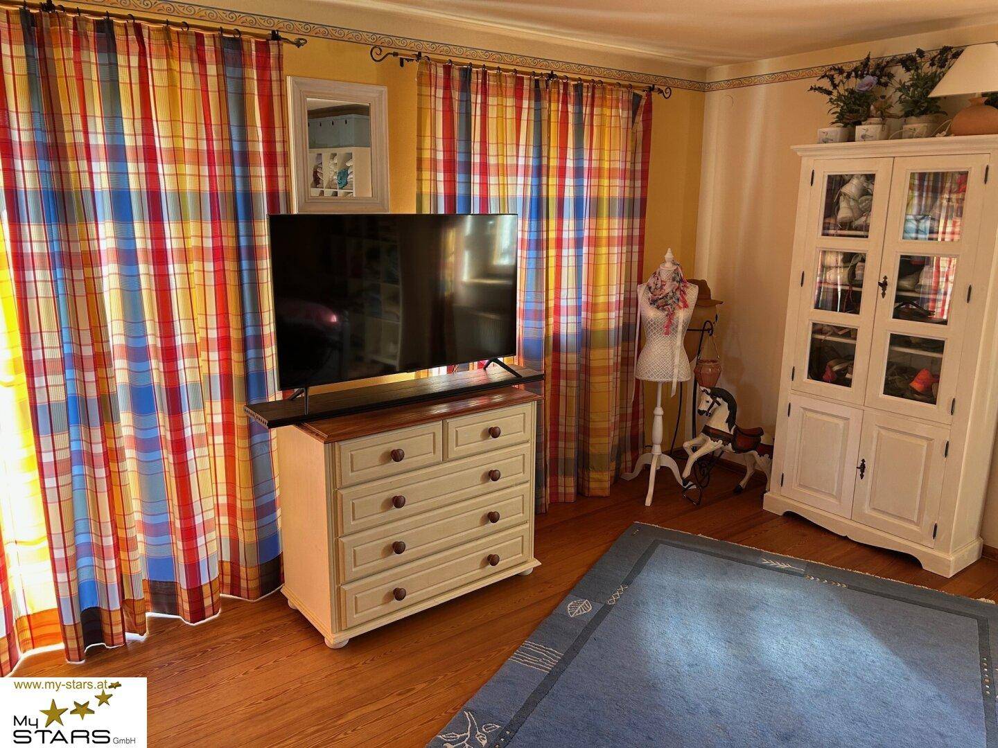Helles Zimmer mit bunten Vorhängen, Kommode und Fernseher, bietet eine freundliche Atmosphäre.