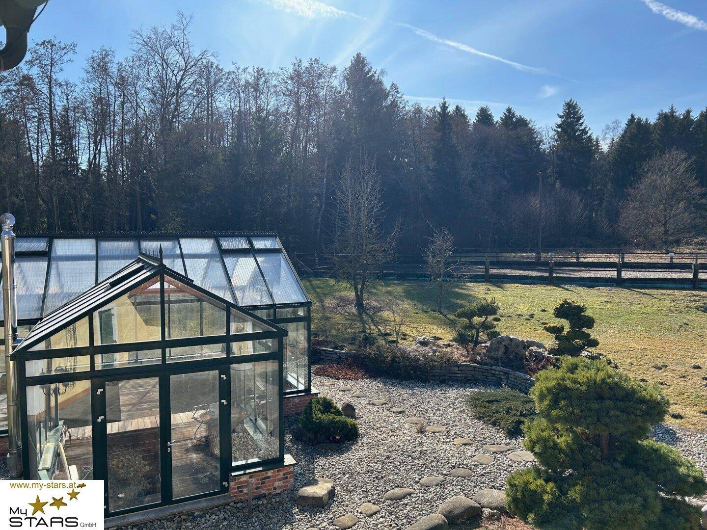 Blick von oben auf ein großes Gewächshaus in einem gepflegten Steingarten mit Zierbäumen.