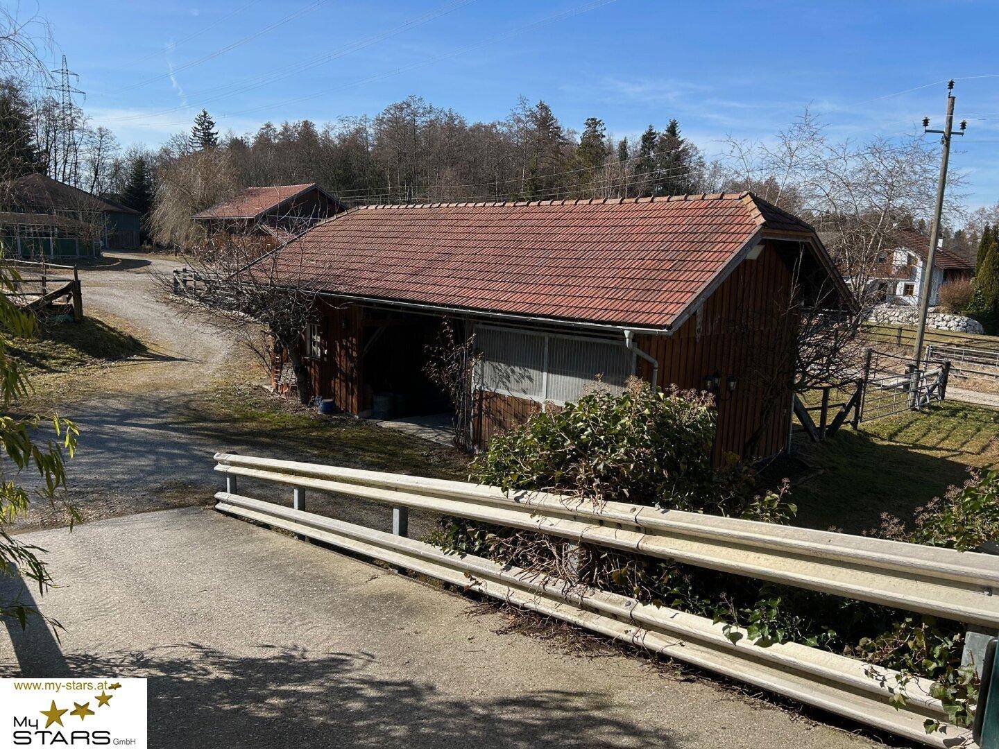 Rustikales Holzgebäude mit Zufahrtsrampe, umgeben von Bäumen und ländlicher Umgebung.