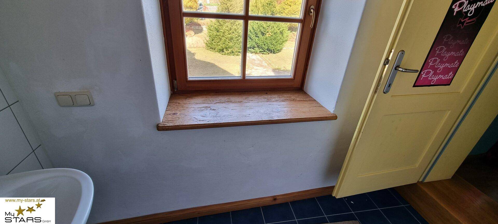 Badezimmerfenster mit Holzrahmen und Blick ins Grüne, über blauen Bodenfliesen.