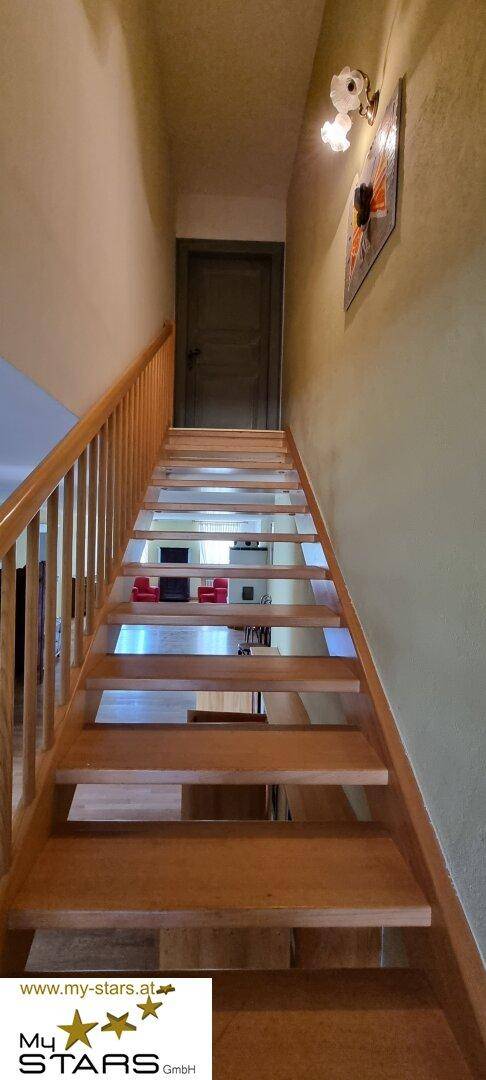 Holztreppe mit Geländer führt nach oben, beleuchtet von einer Wandleuchte.
