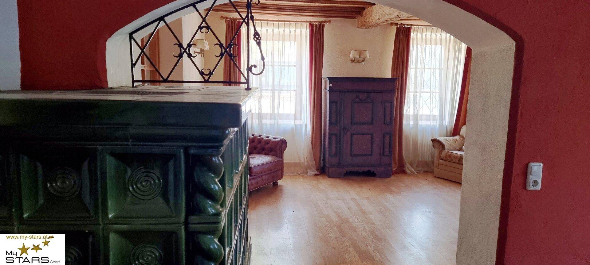 Blick durch einen Bogen auf einen traditionellen Kachelofen und einen hellen Wohnbereich.