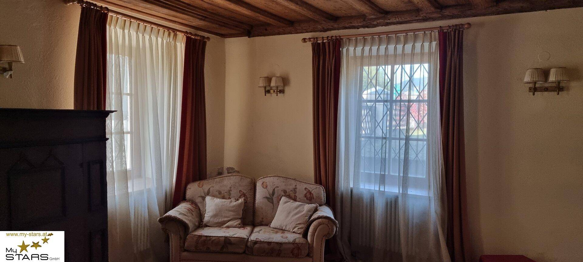 Gemütliches Schlafzimmer mit Holzbalkendecke, zwei Fenstern und traditionellem Sofa.