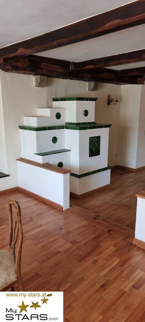 Großer, traditioneller Kachelofen in einem Raum mit Holzboden und Holzbalkendecke.