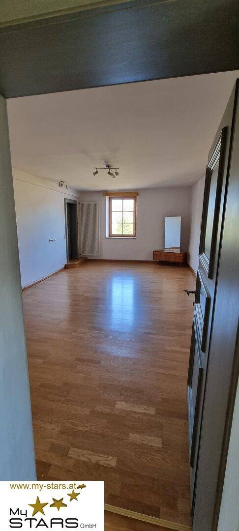 Helles, leeres Zimmer mit Holzboden und einem Fenster, das viel Tageslicht hereinlässt.