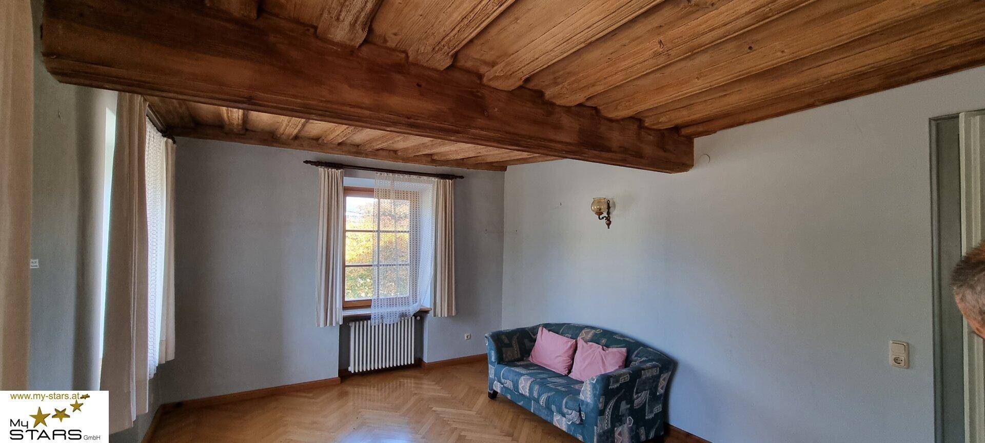 Kleineres Zimmer mit Holzbalkendecke, Parkettboden und einem Sofa am Fenster.