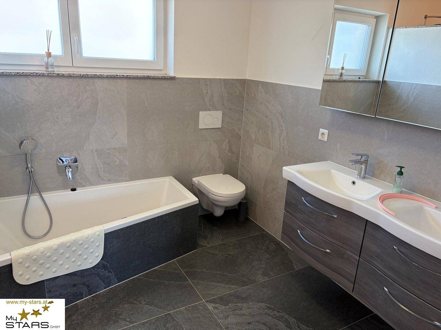 Modernes Badezimmer mit Badewanne, Waschtisch und grauen Fliesen an Wänden und Boden.