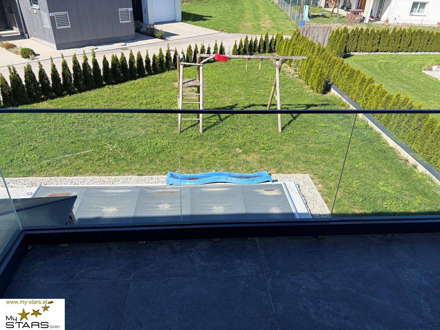 Balkon mit Glasgeländer und Blick auf den Garten mit Schaukel und Poolbereich.