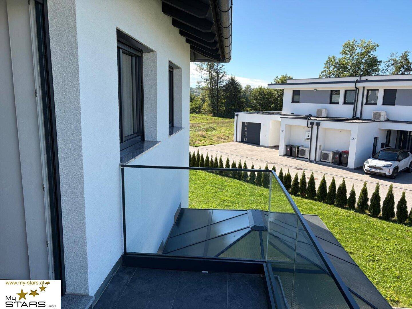 Balkon mit Glasgeländer und Blick auf die grüne Umgebung und Nachbargebäude.