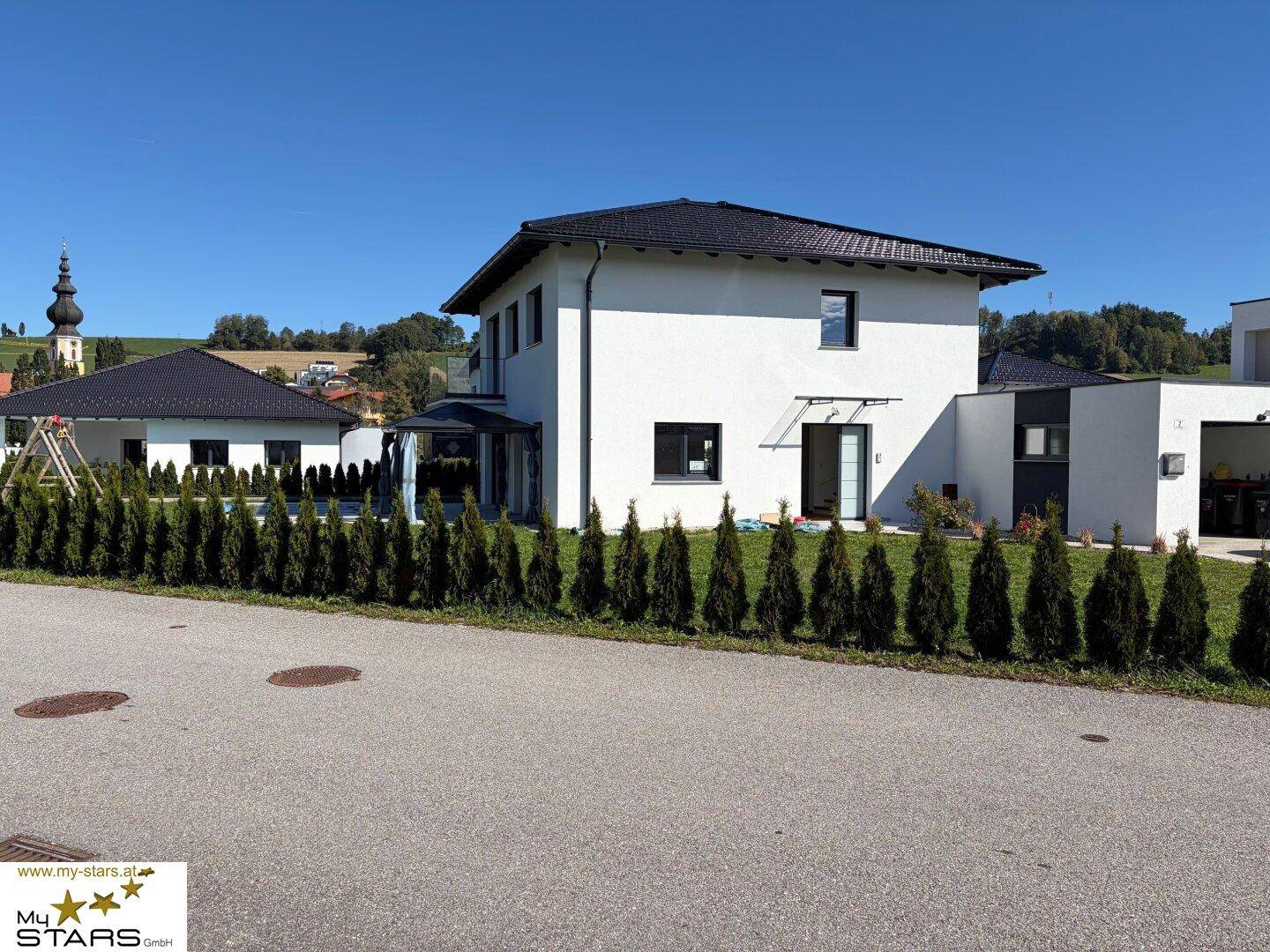 Modernes Einfamilienhaus mit Garten, Hecke und Blick auf die Kirche im Hintergrund.