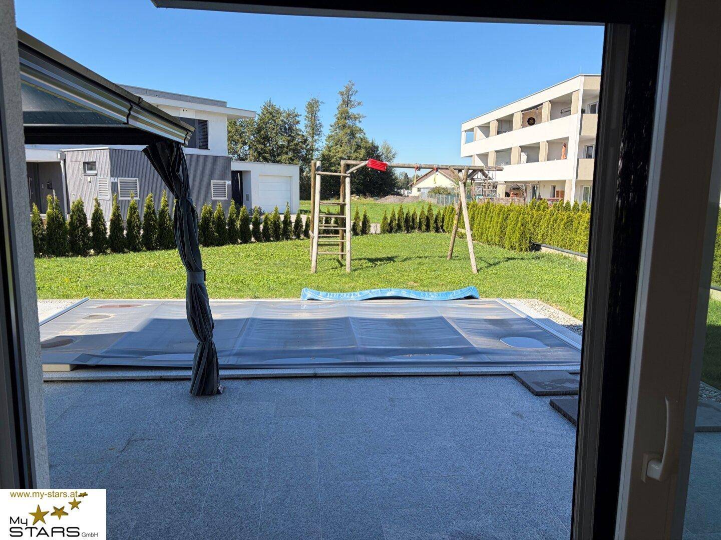 Blick aus dem Wohnzimmer auf die Terrasse, den Garten mit Schaukel und den Poolbereich.