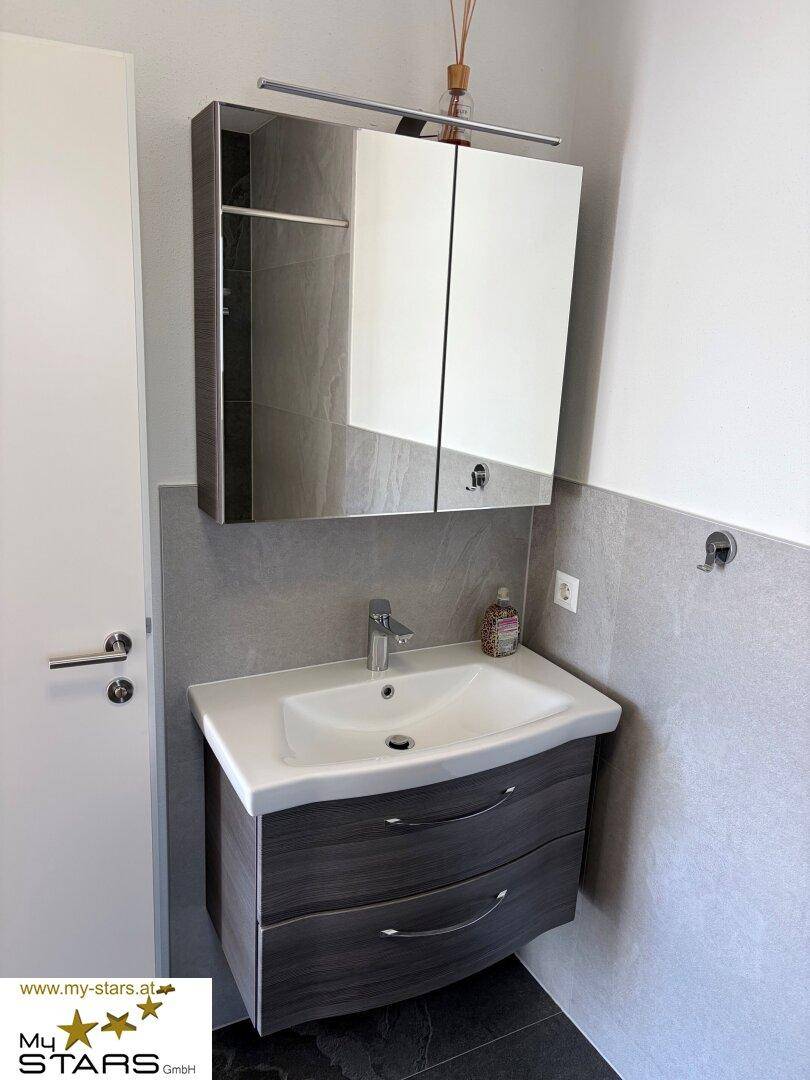 Modernes Badezimmer mit Waschtischunterschrank, Spiegel und hellgrauen Wandfliesen.