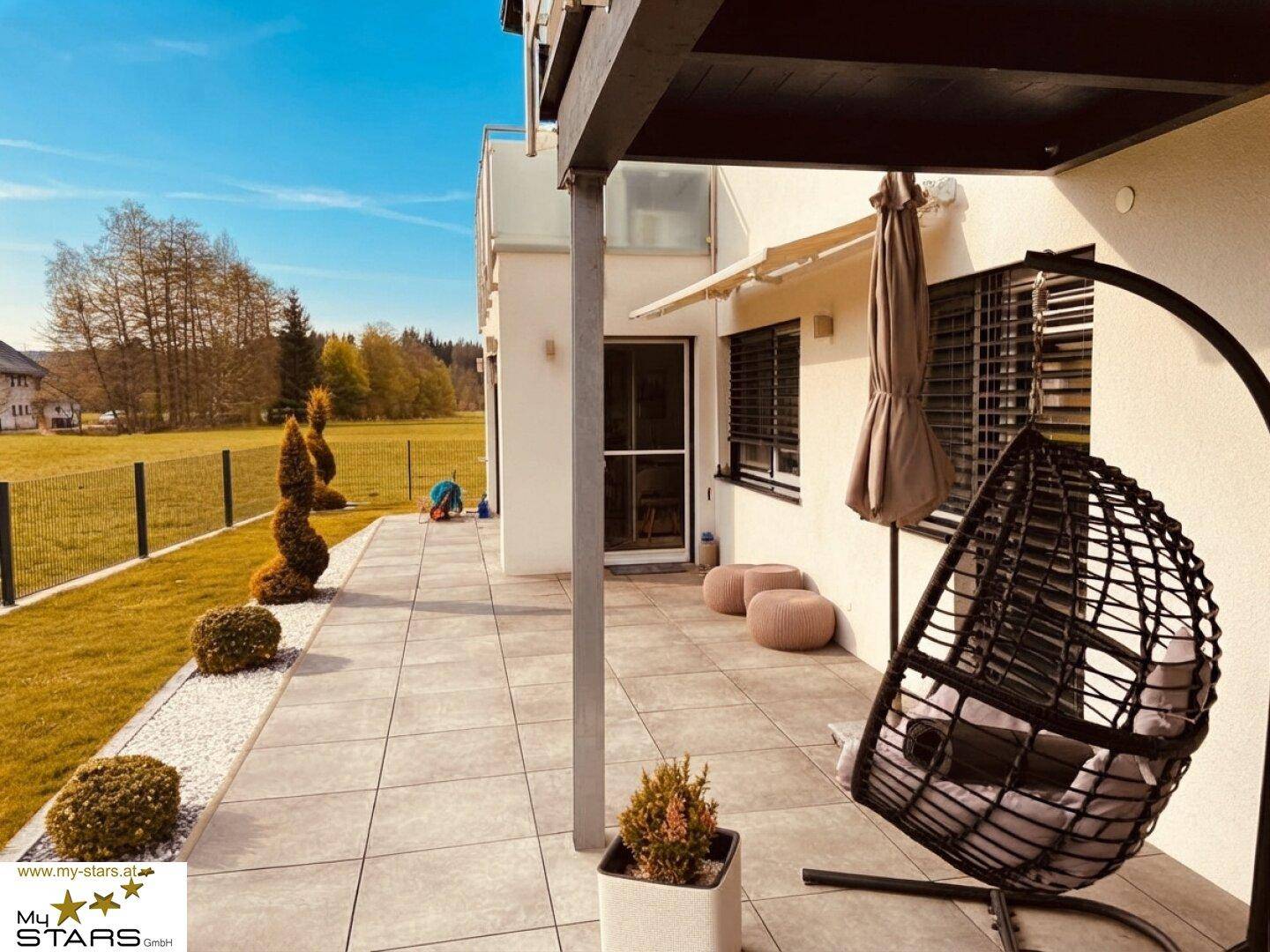 Sonniger Terrassenbereich mit Hängesessel, Liegestühlen und Blick auf den weitläufigen Garten.
