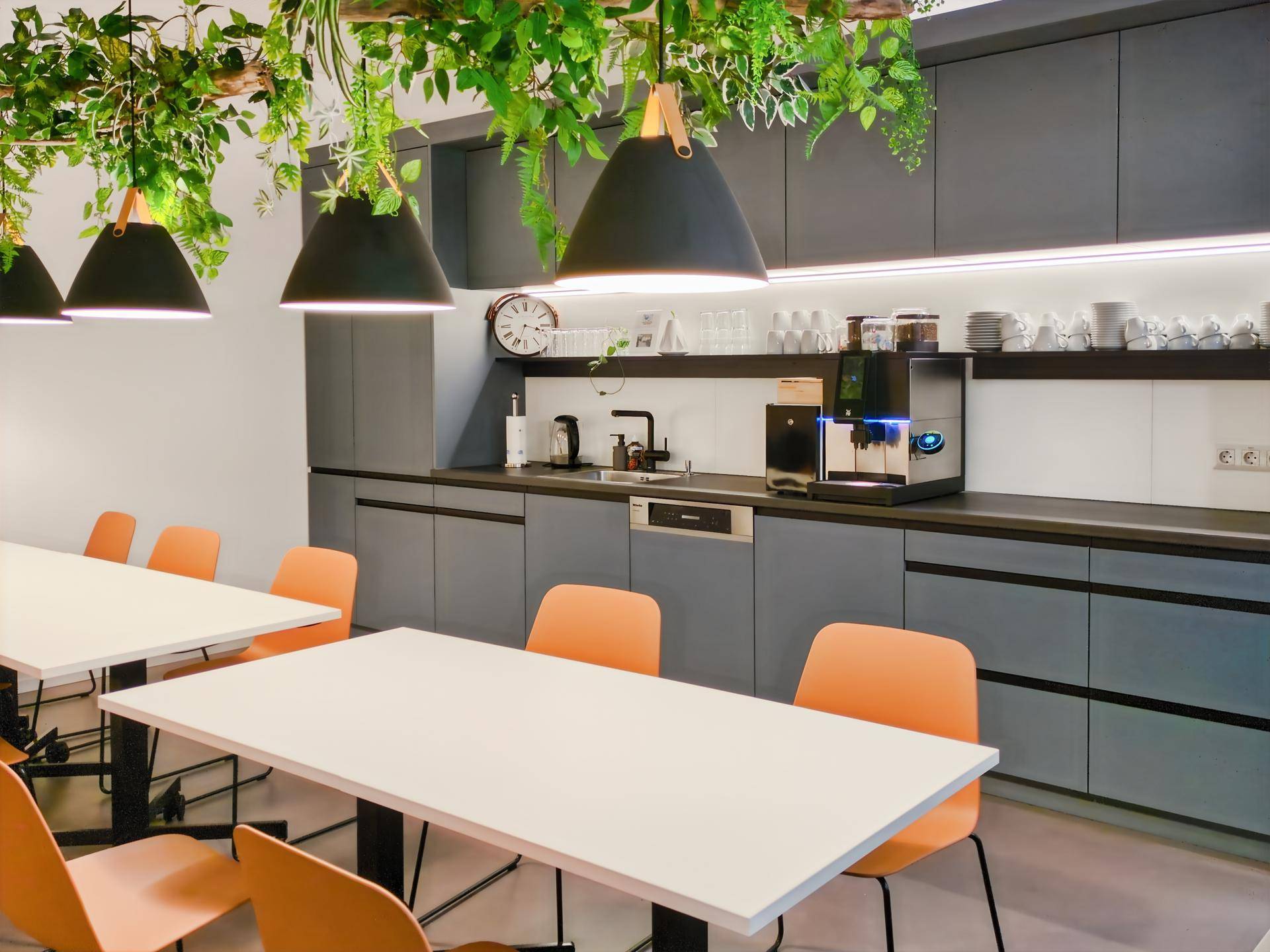 Moderne Teeküche mit grauen Schränken, Kaffeemaschine und Esstischen mit orangefarbenen Stühlen.