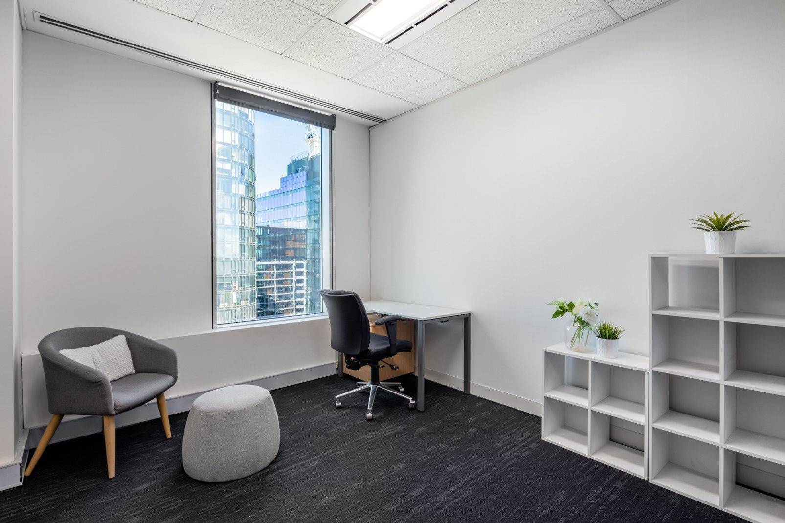 Kleines, helles Büro mit Schreibtisch, Sitzgelegenheiten und Blick auf die Stadt durch ein großes Fenster.
