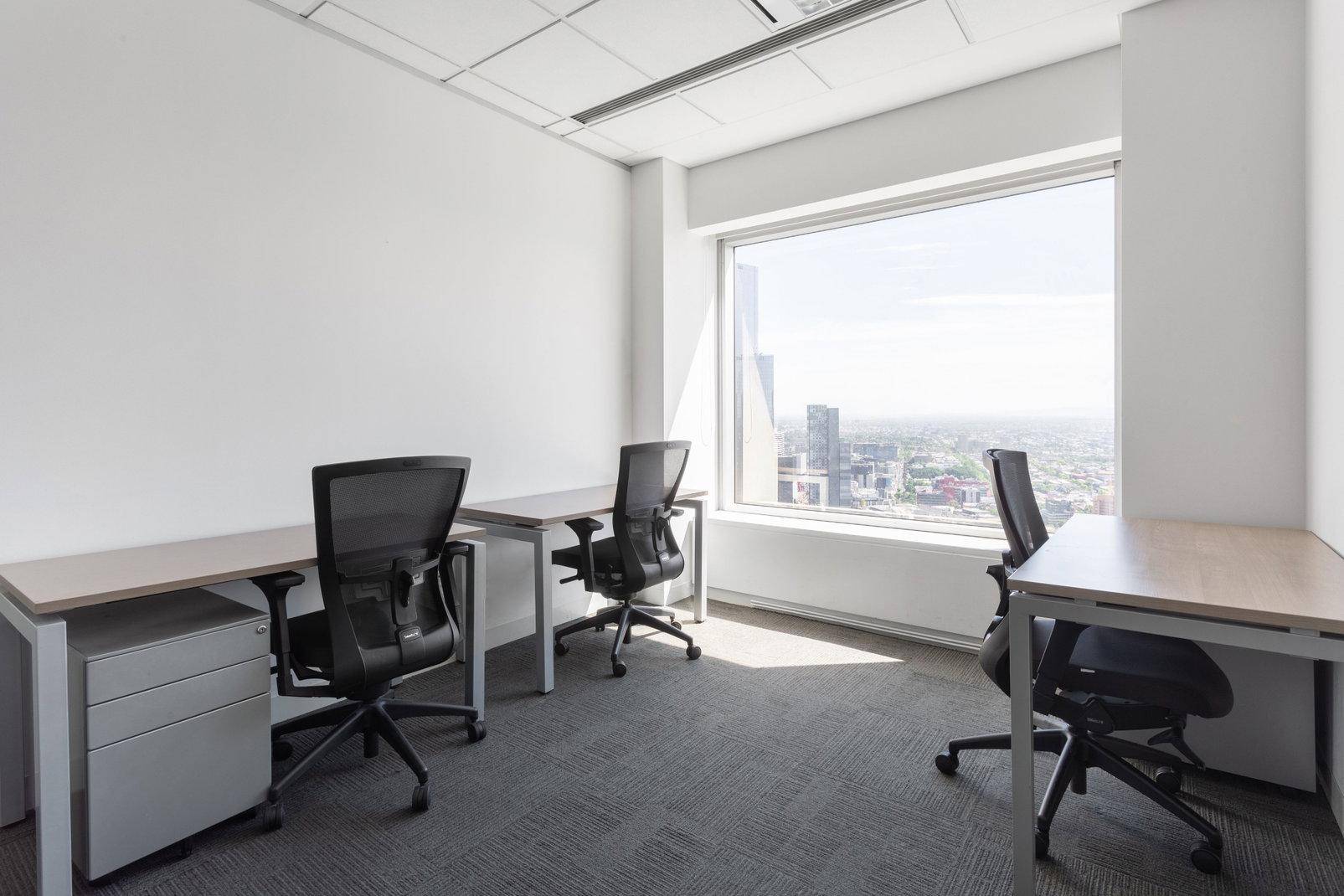 Helles Büro mit mehreren Arbeitsplätzen, Teppichboden und einem großen Fenster mit Stadtblick.