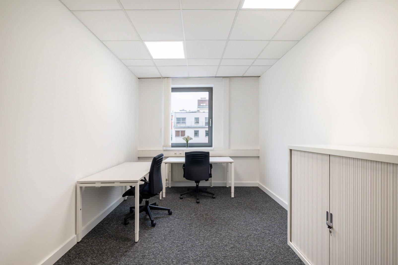 Kleines, helles Büro mit zwei Arbeitsplätzen, Teppichboden und Fensterblick auf die Stadt.
