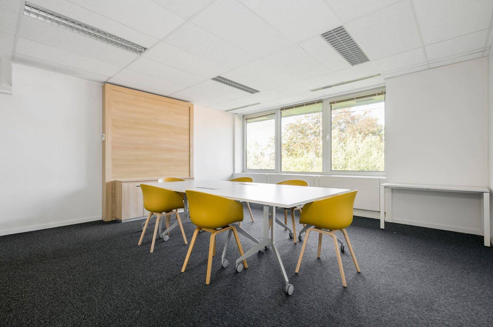 Funktionaler Konferenzraum mit großem Tisch, gelben Stühlen und Teppichboden.