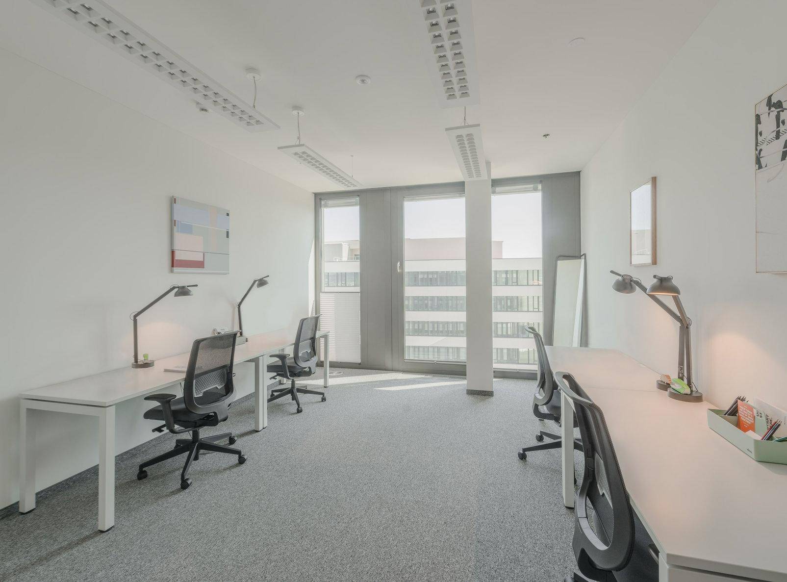 Helles Büro mit mehreren Arbeitsplätzen, Schreibtischen und großen Fenstern mit Blick auf andere Gebäude.