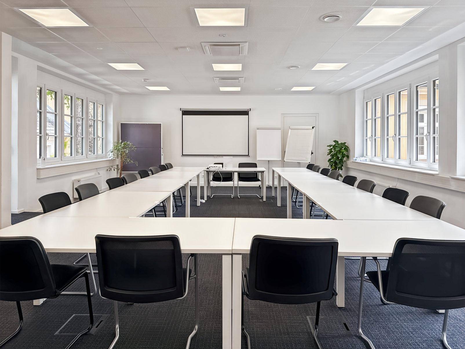 Großer Konferenzraum mit U-förmiger Tischordnung, Projektionsleinwand und vielen Fenstern für Tageslicht.