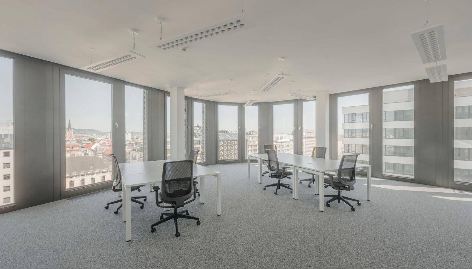 Heller Großraumbüro mit mehreren Schreibtischen, Bürostühlen und großen Fenstern mit Stadtblick.