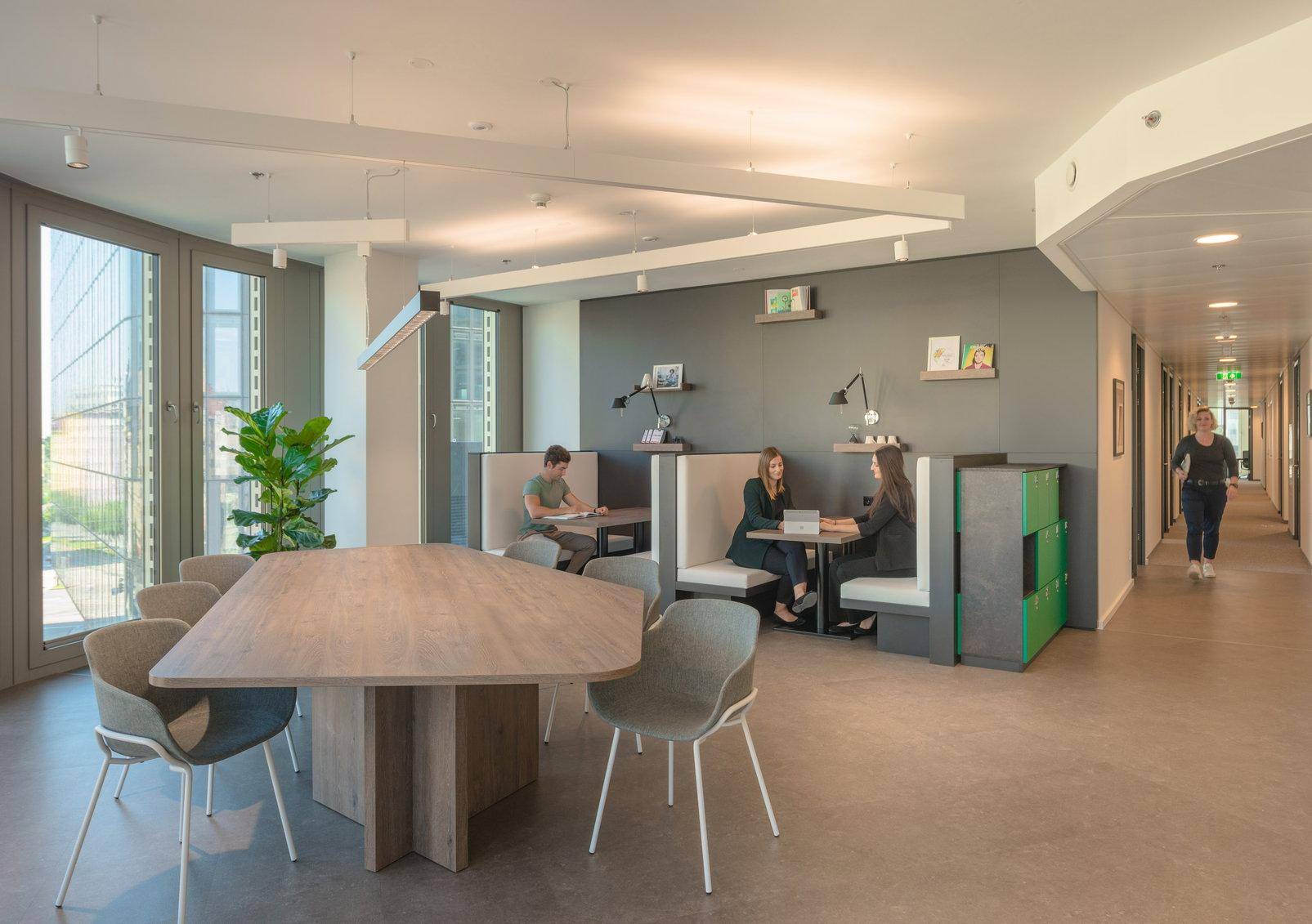 Moderner Gemeinschaftsbereich im Büro mit verschiedenen Sitzgelegenheiten, Besprechungsnischen und großen Fenstern.