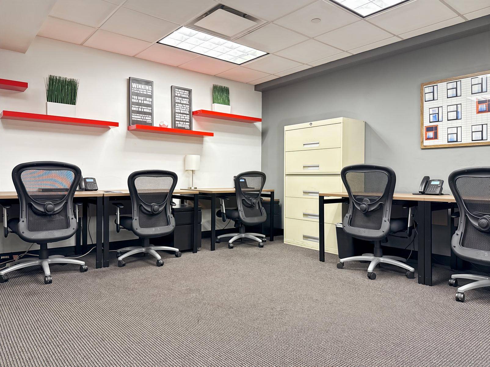 Funktionales Büro mit mehreren Schreibtischen, grauen Wänden und roten Akzentregalen, ausgestattet für mehrere Mitarbeiter.