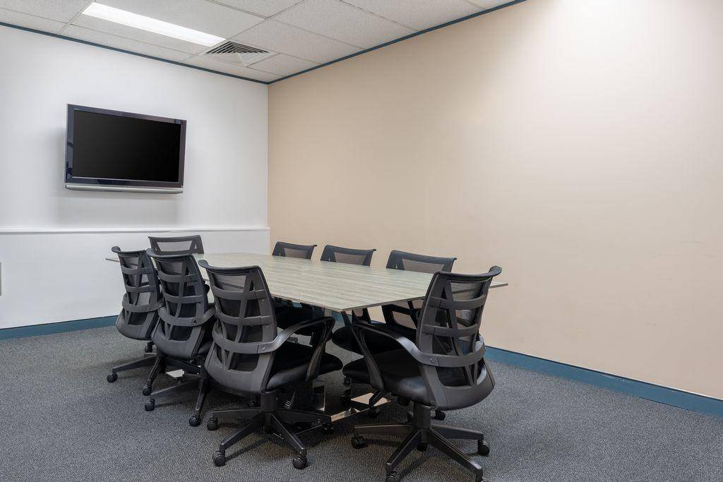 Konferenzraum mit großem Tisch, mehreren Stühlen und einem an der Wand montierten Flachbildschirm.